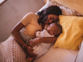 A baby up against his mother and the bed is upagainst the wall.